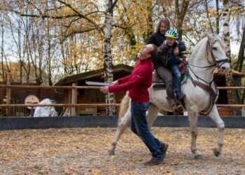 A man in a red sweater playfully pretends to be struck by a toy sword held by a child on horseback. The child, wearing a blue helmet, sits on a white horse behind an adult in a circular sand arena. The background features autumn trees with yellow leaves and a small wooden shed.