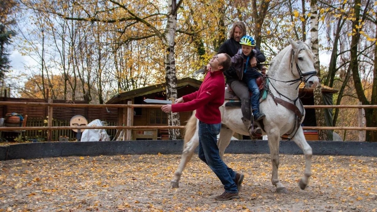 A man in a red sweater playfully pretends to be struck by a toy sword held by a child on horseback. The child, wearing a blue helmet, sits on a white horse behind an adult in a circular sand arena. The background features autumn trees with yellow leaves and a small wooden shed.
