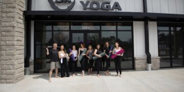 A group of eight people, seven women and one man, stand smiling in front of the entrance to Sol Hot Yoga. They are all barefoot and holding rolled-up yoga mats in various colors like purple, green, and pink. The studio has a modern white and stone exterior with a large black "Sol HOT YOGA" sign above the glass doors.