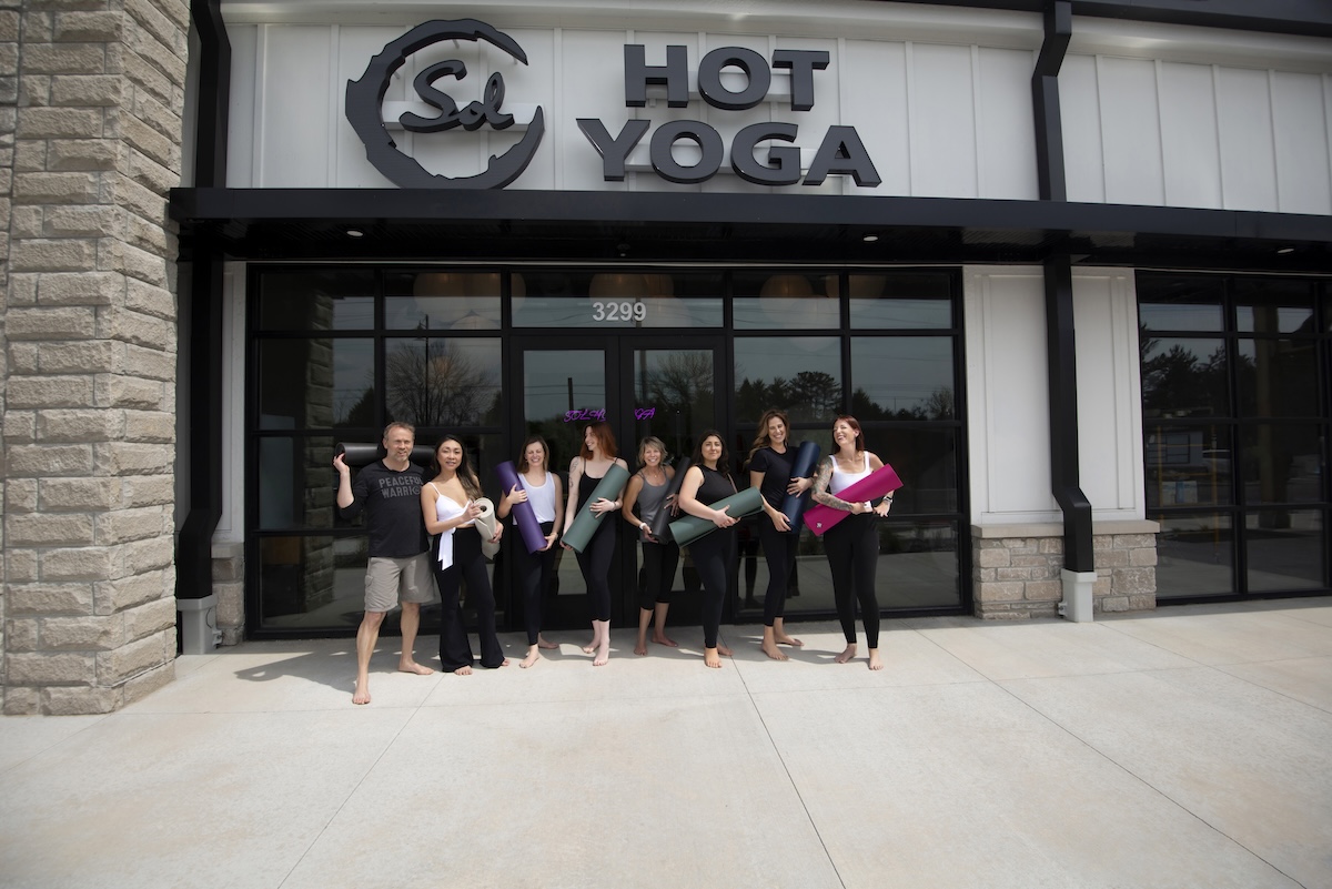 A group of eight people, seven women and one man, stand smiling in front of the entrance to Sol Hot Yoga. They are all barefoot and holding rolled-up yoga mats in various colors like purple, green, and pink. The studio has a modern white and stone exterior with a large black "Sol HOT YOGA" sign above the glass doors.