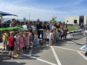 A large crowd of young children and adults gather outdoors for a ribbon-cutting ceremony in a paved area next to a playground fence. Adults hold a long green ribbon while children look on, with a commercial building and rows of folding chairs visible in the background under a blue sky.