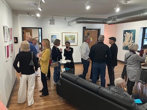 A group of people chat and mingle inside a brightly lit art gallery with white walls and hardwood floors. Framed artwork and paintings hang on the walls around the room, and a black leather sofa sits in the foreground.