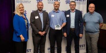 A group of five professionals stands in front of an "Invest Hamilton County, IN" backdrop. From left to right: a woman in a bright blue blazer, a man in a grey suit, a man in a light blue shirt holding a glass award, a man in a navy blazer and glasses, and a man in a blue polo shirt and grey trousers. They are all smiling at the camera during an indoor event.