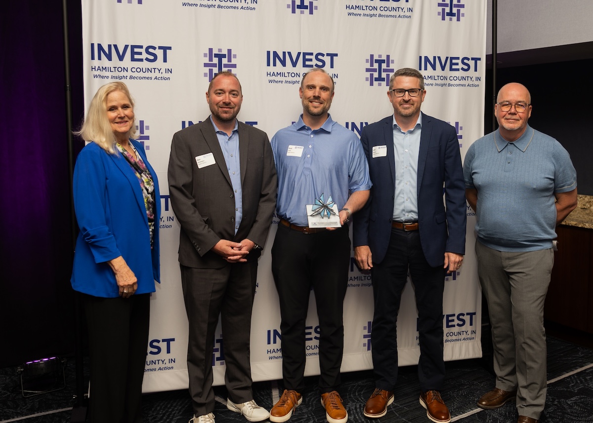 A group of five professionals stands in front of an "Invest Hamilton County, IN" backdrop. From left to right: a woman in a bright blue blazer, a man in a grey suit, a man in a light blue shirt holding a glass award, a man in a navy blazer and glasses, and a man in a blue polo shirt and grey trousers. They are all smiling at the camera during an indoor event.
