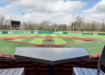 A view from behind home plate at a baseball stadium, looking out over a green and brown turf field. In the foreground, a dark gray table shaped like home plate sits between two empty stadium chairs with wooden slats. The background features the outfield grass, a scoreboard, outfield walls with advertisements, and a line of green trees under a blue sky with white clouds.