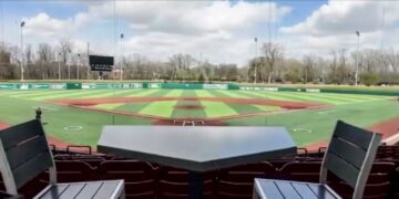 A view from behind home plate at a baseball stadium, looking out over a green and brown turf field. In the foreground, a dark gray table shaped like home plate sits between two empty stadium chairs with wooden slats. The background features the outfield grass, a scoreboard, outfield walls with advertisements, and a line of green trees under a blue sky with white clouds.