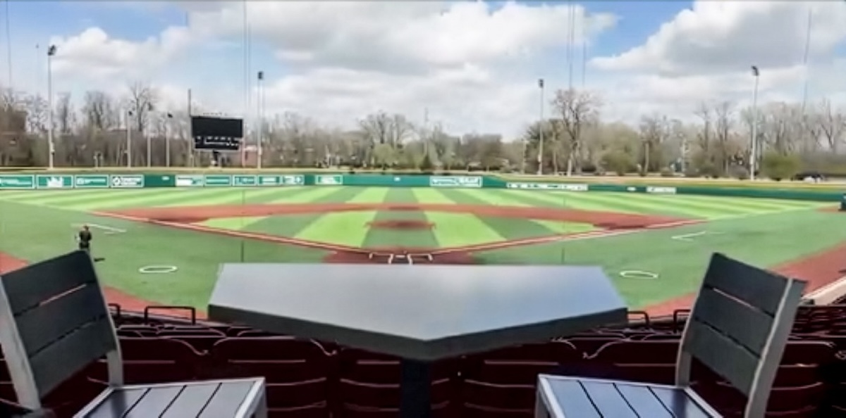 A view from behind home plate at a baseball stadium, looking out over a green and brown turf field. In the foreground, a dark gray table shaped like home plate sits between two empty stadium chairs with wooden slats. The background features the outfield grass, a scoreboard, outfield walls with advertisements, and a line of green trees under a blue sky with white clouds.