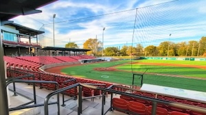 A wide-angle, high-angle view of a baseball stadium from the upper stands behind a protective net. Sections of empty red stadium seats curve along the third-base line toward a multi-story grandstand and press box. The artificial turf field features vibrant green grass and brown dirt paths, bordered by a green outfield wall, tall light poles, and autumn trees under a bright blue sky with wispy clouds.