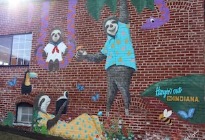 A whimsical mural painted on a red brick wall titled "Hangin' out in INDIANA." The artwork features three sloths: one hanging from a tree branch in a blue tropical shirt, another sitting on a swing in a white outfit, and a third lying on the grass reading a book. The scene is filled with colorful details like a purple snake, a toucan, butterflies, and a bumblebee.