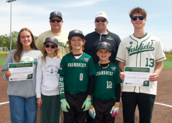 A group of eight people, including four children and four adults, standing on a baseball dirt infield under a clear sky. Two young adults on the ends each hold a "Tucker Swain Memorial Scholarship" certificate. The children in the center wear green "Shamrocks" baseball jerseys and hats. Everyone is smiling for the camera, with a green baseball field and trees in the background.