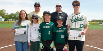 A group of eight people, including four children and four adults, standing on a baseball dirt infield under a clear sky. Two young adults on the ends each hold a "Tucker Swain Memorial Scholarship" certificate. The children in the center wear green "Shamrocks" baseball jerseys and hats. Everyone is smiling for the camera, with a green baseball field and trees in the background.