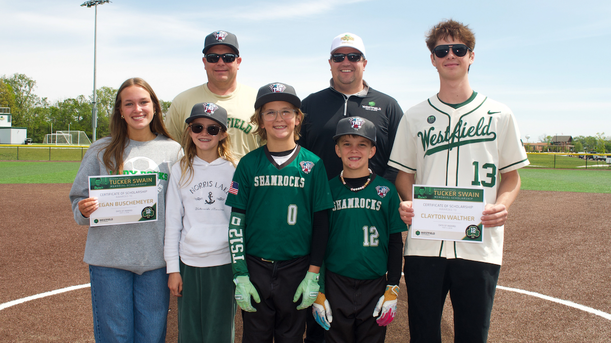 A group of eight people, including four children and four adults, standing on a baseball dirt infield under a clear sky. Two young adults on the ends each hold a "Tucker Swain Memorial Scholarship" certificate. The children in the center wear green "Shamrocks" baseball jerseys and hats. Everyone is smiling for the camera, with a green baseball field and trees in the background.