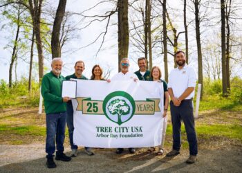 A group of seven people stands in a wooded area, smiling and holding a large white "Tree City USA" banner that celebrates "25 Years." The man on the far left holds a framed certificate. They are outdoors on a sunny day with tall, slender trees and bright green spring foliage in the background.