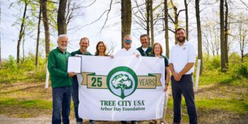 A group of seven people stands in a wooded area, smiling and holding a large white "Tree City USA" banner that celebrates "25 Years." The man on the far left holds a framed certificate. They are outdoors on a sunny day with tall, slender trees and bright green spring foliage in the background.