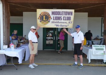 Two older men in white shirts and shorts wave while standing outdoors underneath a large banner that reads "Middletown Lions Club, Middletown, KY, 'We Serve.'" Tables covered in white tablecloths with various items are visible on both sides behind them.