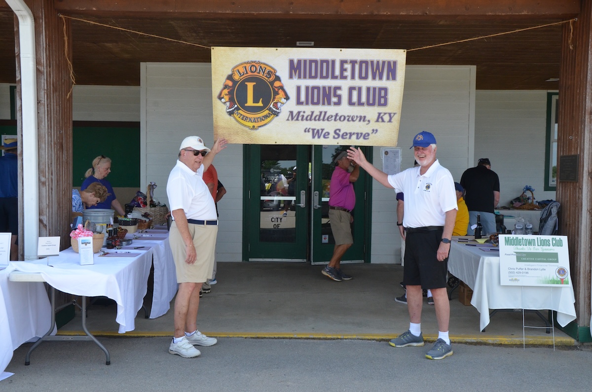 Two older men in white shirts and shorts wave while standing outdoors underneath a large banner that reads "Middletown Lions Club, Middletown, KY, 'We Serve.'" Tables covered in white tablecloths with various items are visible on both sides behind them.