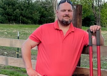 A full-length outdoor portrait of a man with a goatee and sunglasses resting on his head. He wears a bright coral-red polo shirt, a dark leather belt, and stained khaki pants. He stands with his right hand on his hip and his left arm resting on a tall, red-handled post hole digger tool. He is leaning against a rustic wooden horizontal rail fence. In the background, an open grassy field extends toward a dense line of green pine trees under a pale blue sky with light clouds.