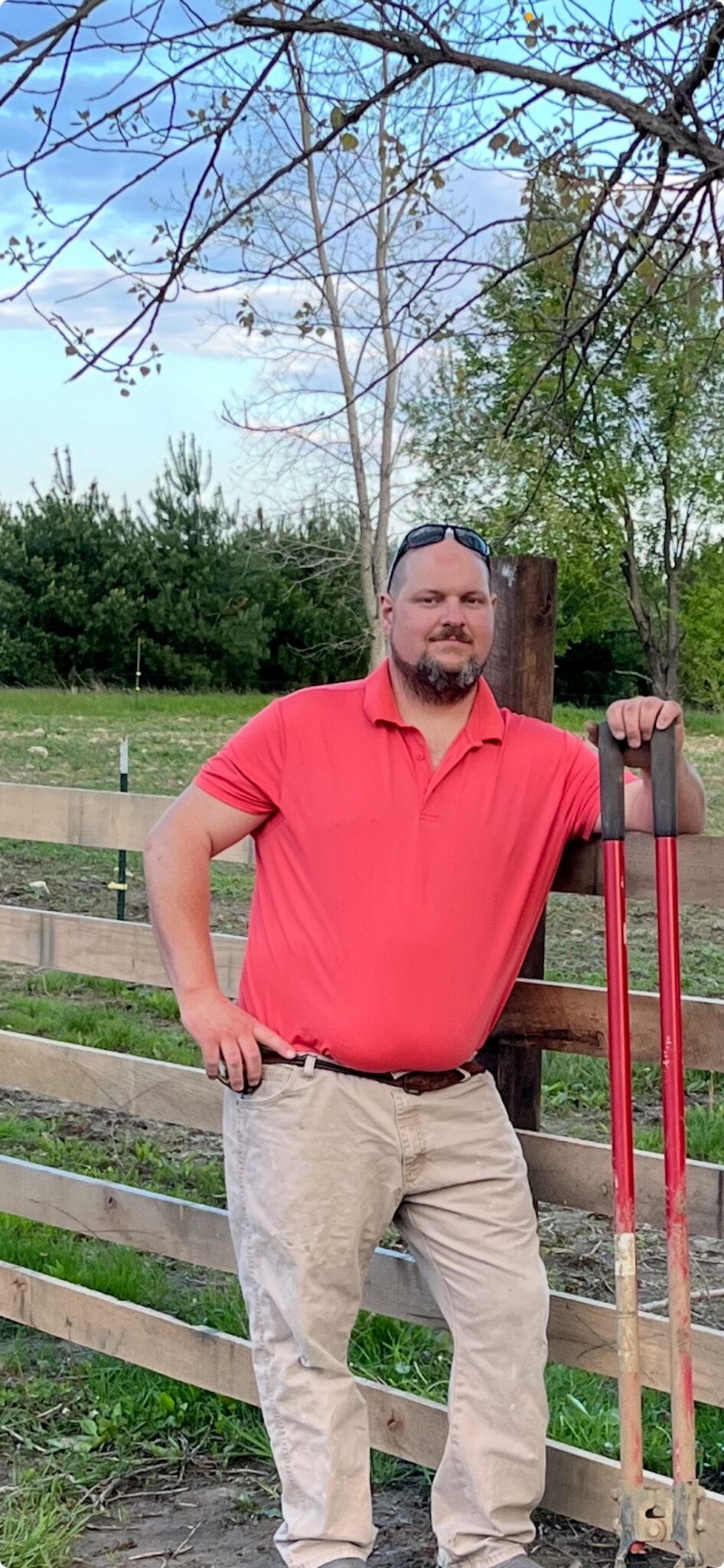A full-length outdoor portrait of a man with a goatee and sunglasses resting on his head. He wears a bright coral-red polo shirt, a dark leather belt, and stained khaki pants. He stands with his right hand on his hip and his left arm resting on a tall, red-handled post hole digger tool. He is leaning against a rustic wooden horizontal rail fence. In the background, an open grassy field extends toward a dense line of green pine trees under a pale blue sky with light clouds.