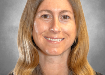 A professional headshot of a woman with long, light brown wavy hair and a pleasant smile. She is wearing a black crew-neck shirt. The portrait is set against a neutral, mottled grey studio background.