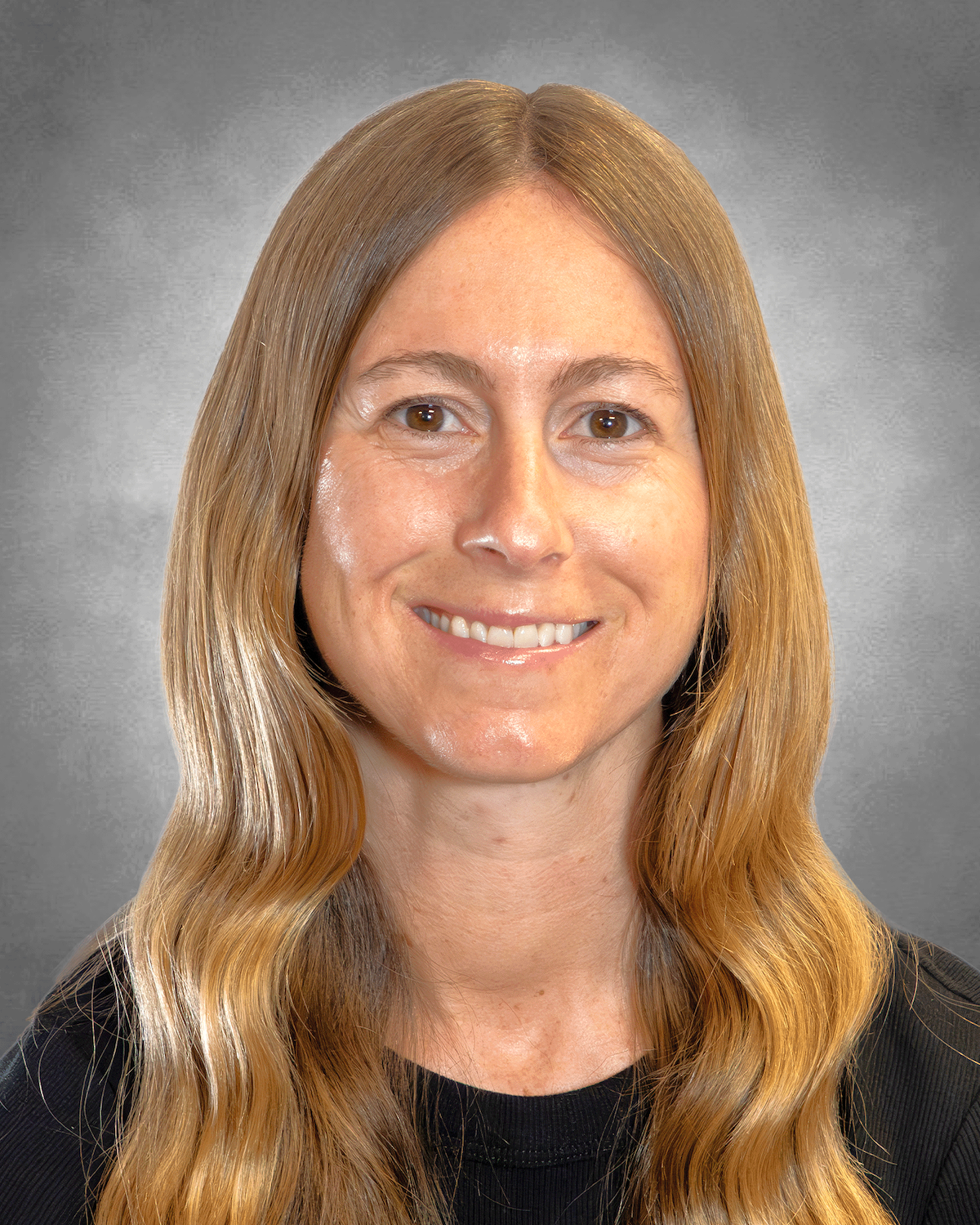 A professional headshot of a woman with long, light brown wavy hair and a pleasant smile. She is wearing a black crew-neck shirt. The portrait is set against a neutral, mottled grey studio background.