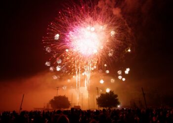 A wide shot of a large, vibrant pink and white firework exploding in the night sky over a dark crowd of people. The firework is the central focus, with smaller sparks trailing below it. Silhouetted trees and stadium lights are visible in the mid-ground. The bottom of the frame shows the backs of many people gathered to watch the display.