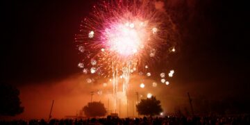 A wide shot of a large, vibrant pink and white firework exploding in the night sky over a dark crowd of people. The firework is the central focus, with smaller sparks trailing below it. Silhouetted trees and stadium lights are visible in the mid-ground. The bottom of the frame shows the backs of many people gathered to watch the display.