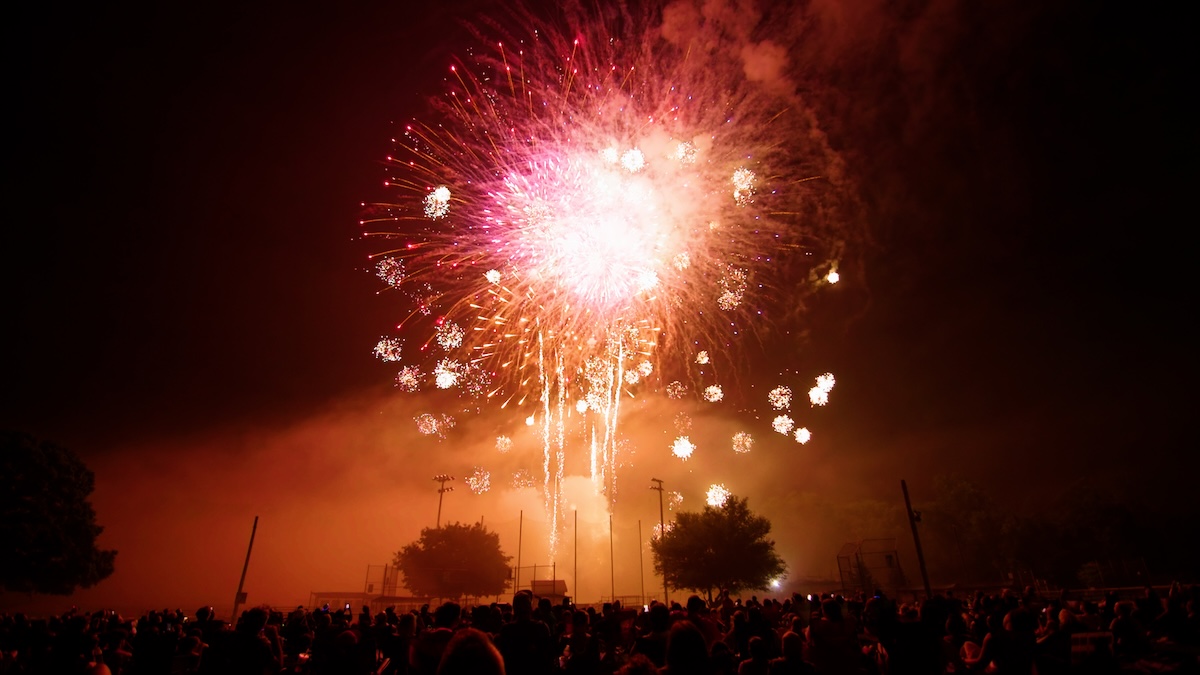 A wide shot of a large, vibrant pink and white firework exploding in the night sky over a dark crowd of people. The firework is the central focus, with smaller sparks trailing below it. Silhouetted trees and stadium lights are visible in the mid-ground. The bottom of the frame shows the backs of many people gathered to watch the display.
