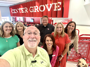 A group of six smiling adults and one yellow Labrador Retriever stand together in a modern school hallway. In the background, a large red wall features the words "CENTER GROVE" in bold white letters. The group includes several women in casual and business-casual attire and one man taking the selfie. One woman on the right holds the dog’s red leash.