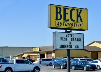 A large yellow and black sign for Beck Automotive stands tall in a busy parking lot. A smaller white marquee below it reads "VOTED BEST GARAGE IN JOHNSON CO." In the background, the automotive service building is visible with several cars parked in front under a clear blue sky.