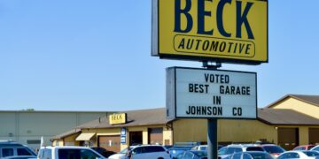 A large yellow and black sign for Beck Automotive stands tall in a busy parking lot. A smaller white marquee below it reads "VOTED BEST GARAGE IN JOHNSON CO." In the background, the automotive service building is visible with several cars parked in front under a clear blue sky.