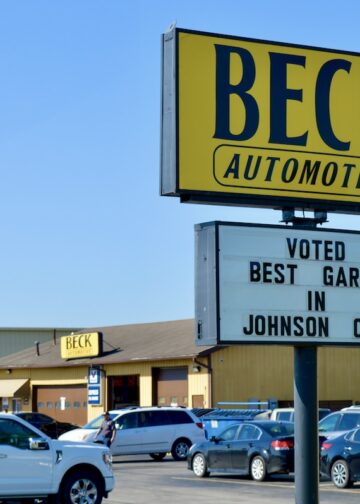 A large yellow and black sign for Beck Automotive stands tall in a busy parking lot. A smaller white marquee below it reads "VOTED BEST GARAGE IN JOHNSON CO." In the background, the automotive service building is visible with several cars parked in front under a clear blue sky.