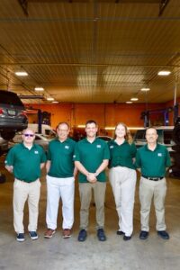 Five employees from Beck Automotive stand in a row inside a large, well-lit service bay. They are wearing matching green polo shirts with the company logo. In the background, several vehicles are being serviced on hydraulic lifts, and the shop's orange walls and industrial lighting are visible.