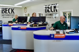 The interior of Beck Automotive, showing three service advisors working behind individual curved white and blue counters. Each advisor is at a computer station in a bright, clean office. Large "BECK AUTOMOTIVE" logos are displayed on the back wall, along with the text "Established in 1988."