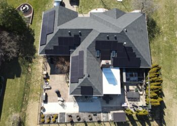 An aerial, top-down drone photograph of a large residential home featuring dark grey roof shingles outfitted with multiple arrays of sleek black solar panels. The surrounding property includes a fenced backyard patio with outdoor furniture, landscaped garden beds, and green grassy lawns.