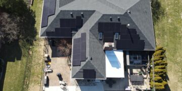 An aerial, top-down drone photograph of a large residential home featuring dark grey roof shingles outfitted with multiple arrays of sleek black solar panels. The surrounding property includes a fenced backyard patio with outdoor furniture, landscaped garden beds, and green grassy lawns.