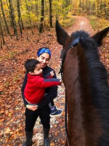A woman in a blue headband and a young child in a red jacket standing on a leaf-covered forest trail. The woman is smiling and holding the child as they interact with a dark brown horse, seen from a high-angle perspective. The background is filled with vibrant autumn trees and fallen orange leaves.
