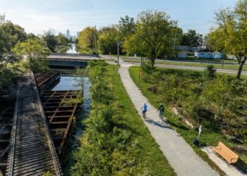 An elevated, wide-angle shot shows two people cycling on a paved trail next to a canal and an old wooden railroad trestle. The cyclists, one in light blue and one in dark clothing, ride away from the camera toward a bridge in the midground. To their left, the weathered wooden slats of the trestle sit above the calm water of the canal. Lush green trees and bushes line the path, and in the far distance, the Indianapolis city skyline is visible under a clear, bright sky.