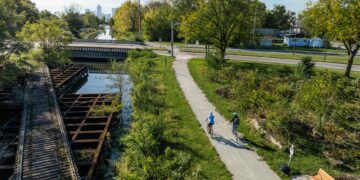 An elevated, wide-angle shot shows two people cycling on a paved trail next to a canal and an old wooden railroad trestle. The cyclists, one in light blue and one in dark clothing, ride away from the camera toward a bridge in the midground. To their left, the weathered wooden slats of the trestle sit above the calm water of the canal. Lush green trees and bushes line the path, and in the far distance, the Indianapolis city skyline is visible under a clear, bright sky.