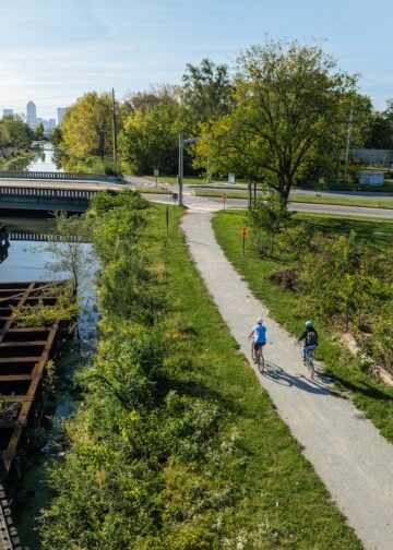 An elevated, wide-angle shot shows two people cycling on a paved trail next to a canal and an old wooden railroad trestle. The cyclists, one in light blue and one in dark clothing, ride away from the camera toward a bridge in the midground. To their left, the weathered wooden slats of the trestle sit above the calm water of the canal. Lush green trees and bushes line the path, and in the far distance, the Indianapolis city skyline is visible under a clear, bright sky.