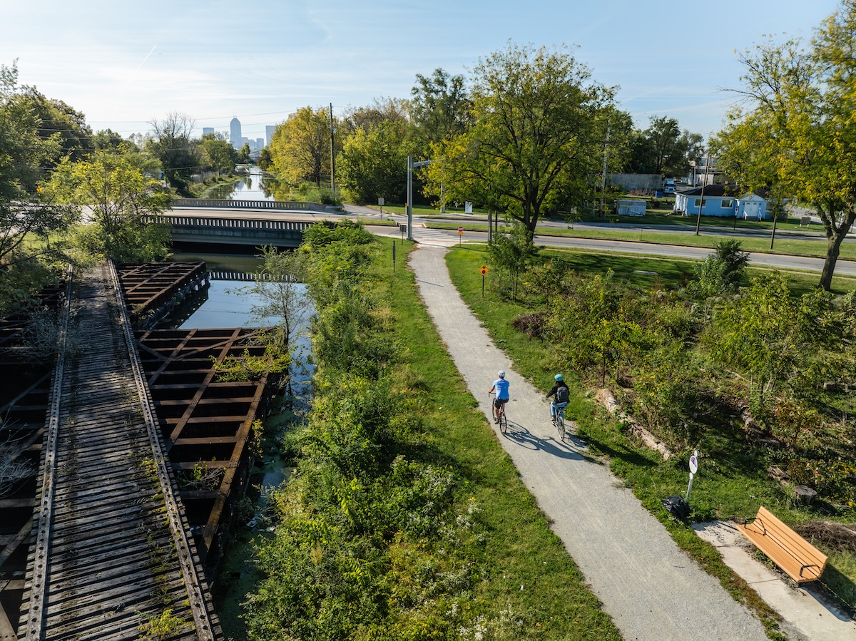 An elevated, wide-angle shot shows two people cycling on a paved trail next to a canal and an old wooden railroad trestle. The cyclists, one in light blue and one in dark clothing, ride away from the camera toward a bridge in the midground. To their left, the weathered wooden slats of the trestle sit above the calm water of the canal. Lush green trees and bushes line the path, and in the far distance, the Indianapolis city skyline is visible under a clear, bright sky.