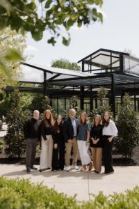 A group of seven people, likely a professional team, stands smiling in front of a modern, black-framed glass structure. The group includes four women and three men, dressed in business-casual attire. They are outdoors on a sunny day, with lush green trees and shrubs framing the scene. The glass building in the background has a unique, geometric roofline with white fabric panels.