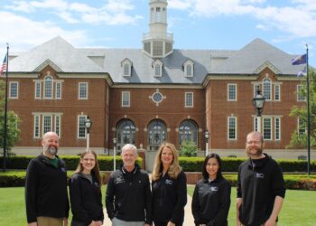 Six smiling people stand in a line on a concrete walkway in front of Carmel City Hall, a large red brick building with a central white clock tower cupola under a bright, partly cloudy sky. The group, consisting of three men and three women, wear matching long-sleeved black shirts featuring a small white logo on the left chest.
