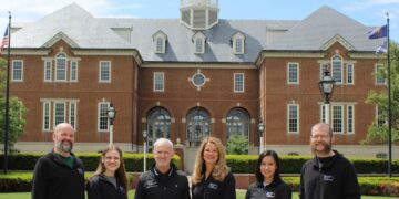 Six smiling people stand in a line on a concrete walkway in front of Carmel City Hall, a large red brick building with a central white clock tower cupola under a bright, partly cloudy sky. The group, consisting of three men and three women, wear matching long-sleeved black shirts featuring a small white logo on the left chest.