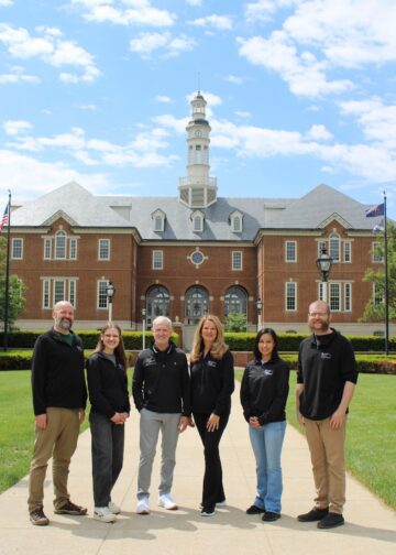 Six smiling people stand in a line on a concrete walkway in front of Carmel City Hall, a large red brick building with a central white clock tower cupola under a bright, partly cloudy sky. The group, consisting of three men and three women, wear matching long-sleeved black shirts featuring a small white logo on the left chest.