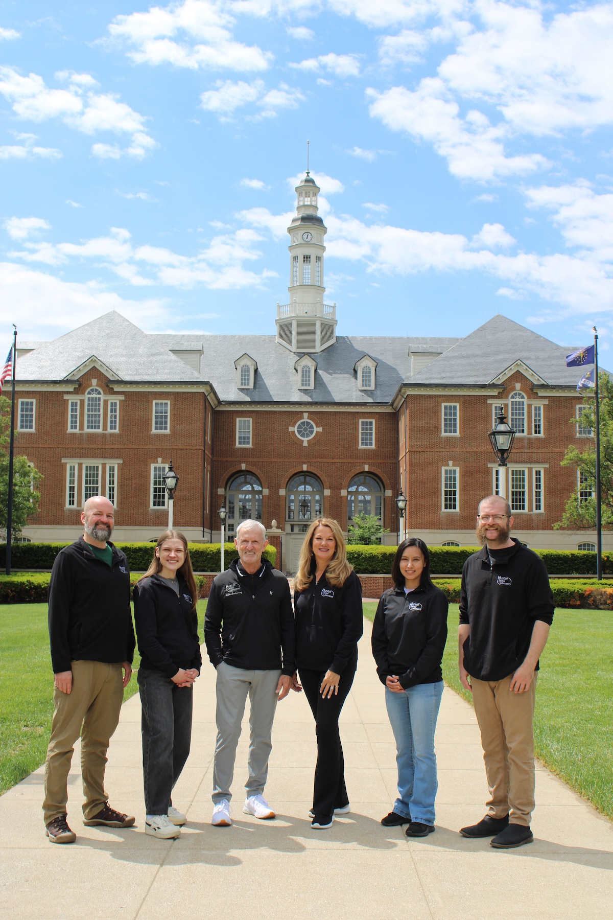 Six smiling people stand in a line on a concrete walkway in front of Carmel City Hall, a large red brick building with a central white clock tower cupola under a bright, partly cloudy sky. The group, consisting of three men and three women, wear matching long-sleeved black shirts featuring a small white logo on the left chest.