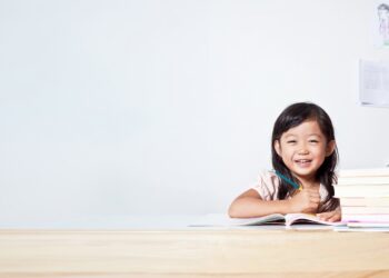 A young girl with dark hair sits at a wooden desk, smiling brightly while holding a pencil over an open notebook. A stack of books sits next to her against a plain white background with ample copy space on the left.