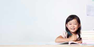 A young girl with dark hair sits at a wooden desk, smiling brightly while holding a pencil over an open notebook. A stack of books sits next to her against a plain white background with ample copy space on the left.