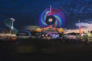 A long-exposure night photograph captures the vibrant energy of the Midway at the Texas State Fair. In the center, a brightly lit entrance arch features the word "MIDWAY" in bold black letters against a glowing yellow and orange background. Behind the sign, a spinning amusement ride creates a large, mesmerizing circular trail of rainbow-colored light that dominates the sky. To the right, another ride appears as a tall, vertical streak of orange light, while the left features a curved blue structure illuminated with neon. The scene is set against a dark blue twilight sky with soft clouds, and the foreground shows the dark, blurred shapes of fairgoers.