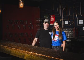 A man and a woman stand together behind a textured, industrial-style bar in a dimly lit space. The man, wearing a black t-shirt and a baseball cap, has his arm around the woman, who is wearing a bright blue "Greek's Pizzeria" t-shirt. The background features a dark wall decorated with a network of copper pipes, small framed quotes, and rows of what look like tap handles or keys. A warm, vintage-style industrial light fixture hangs above them.