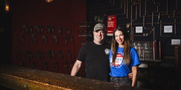 A man and a woman stand together behind a textured, industrial-style bar in a dimly lit space. The man, wearing a black t-shirt and a baseball cap, has his arm around the woman, who is wearing a bright blue "Greek's Pizzeria" t-shirt. The background features a dark wall decorated with a network of copper pipes, small framed quotes, and rows of what look like tap handles or keys. A warm, vintage-style industrial light fixture hangs above them.